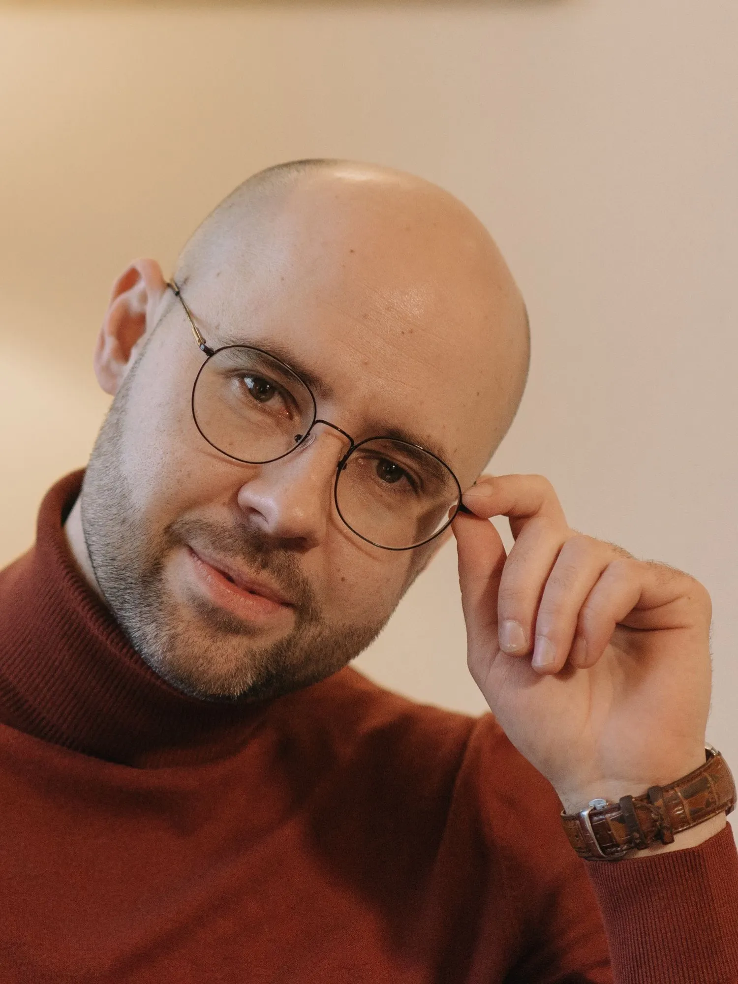Porträt von Lorenz Weidinger mit runder Brille und rotbraunem Rollkragenpullover, im Hintergrund eine Stehlampe.